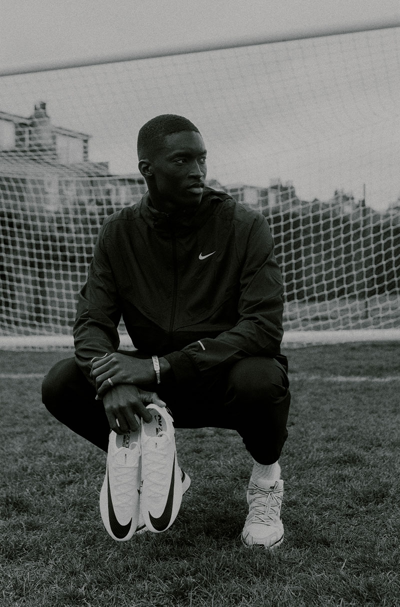 Pape Daouda Diong en noir et blanc sur le terrain d'entraînement de Chelsea Pape Daouda Diong accroupi, tenant une paire de Nike, sur un terrain d'entraînement de Chelsea en noir et blanc.